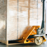 An orange pallet jack lifting a large, wrapped package on a wooden pallet in a warehouse setting- Carlile avoids shipping delays due to the holidays